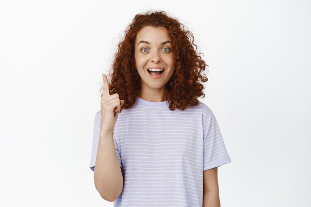 Tengo una idea, gran solución. La chica rizada emocionada tiene un plan, levanta el signo de eureka con el dedo, señala hacia arriba y sonríe, parada en camiseta sobre fondo blanco