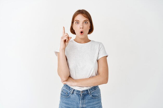 Tengo una idea. Mujer joven emocionada en camiseta, levantando el dedo signo eureka, tiene plan, sugiriendo algo, de pie en la pared blanca