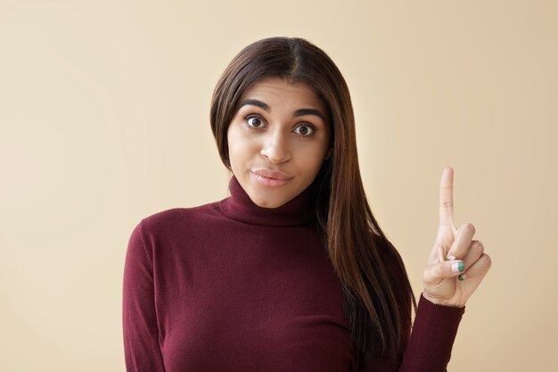 ¡Tengo una idea! Vista recortada de una atractiva joven emocional de piel oscura haciendo una expresión facial divertida y apuntando con el dedo índice hacia arriba para llamar su atención, con una mirada de sorpresa y emoción