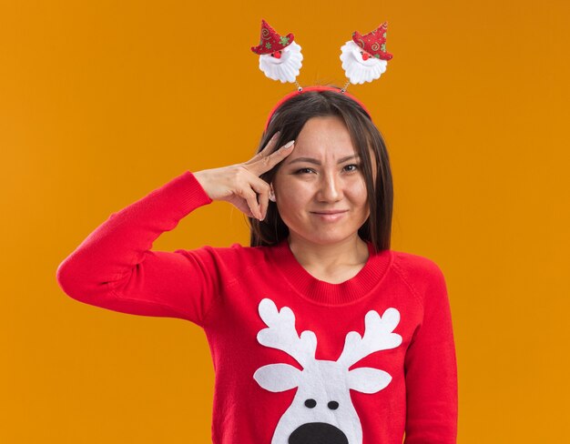 Tensa joven asiática vistiendo aro de pelo de navidad con suéter poniendo los dedos en la cabeza aislada en la pared naranja