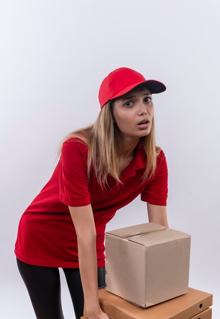 Tensa joven repartidora vestida con uniforme rojo y gorra tratando de sostener muchas cajas aisladas en blanco