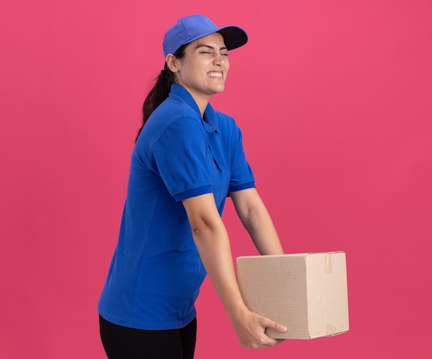 Tensa con los ojos cerrados joven repartidora vistiendo uniforme con gorra sosteniendo la caja aislada en la pared rosa