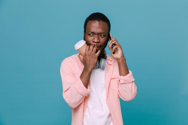 tense habla por teléfono sosteniendo una taza de café joven afroamericano con auriculares en el cuello aislado de fondo azul