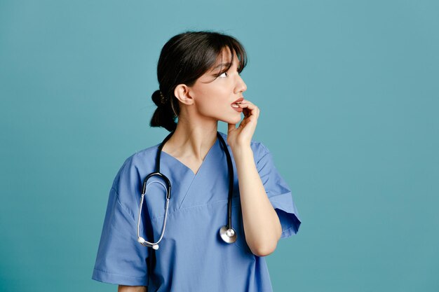 Tenso agarró la barbilla joven doctora vistiendo uniforme fith estetoscopio aislado sobre fondo azul.