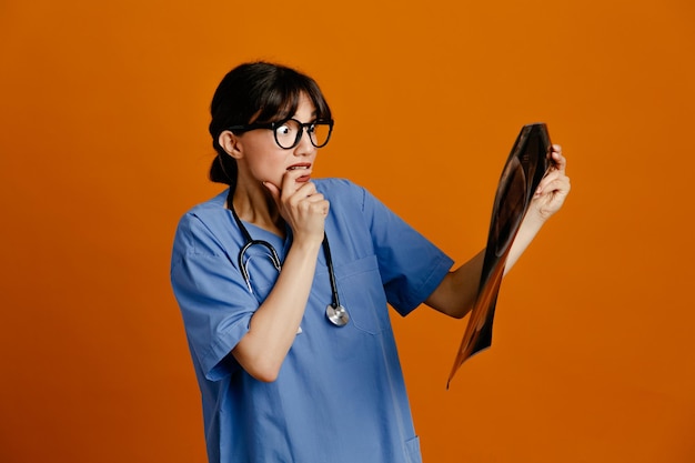 Tenso agarró la barbilla sosteniendo rayos X joven doctora vistiendo uniforme fith estetoscopio aislado sobre fondo naranja