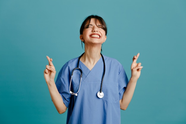 Tenso cruzar los dedos joven doctora vistiendo uniforme fith estetoscopio aislado sobre fondo azul.