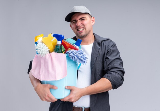 Tenso joven guapo chico de limpieza con camiseta y gorra sosteniendo un balde de herramientas de limpieza aislado en la pared blanca