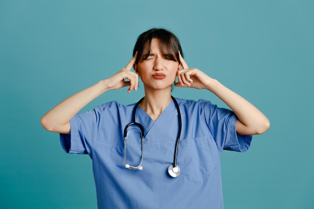 Tenso poner el dedo en la cabeza joven doctora vistiendo uniforme fith estetoscopio aislado sobre fondo azul.