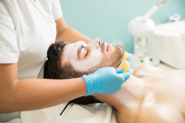 Terapeuta femenina lavando la cara de un cliente masculino mientras le da un tratamiento facial en un spa