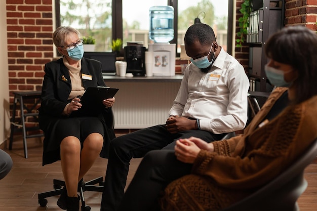 Terapeuta mujer hablando con hombre afroamericano en terapia de grupo de apoyo, sentado en círculo. Personas en una reunión discutiendo problemas de salud mental y adicción al alcohol, usando mascarillas.