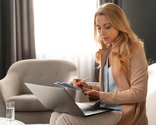 Terapeuta de mujer de tiro medio tomando notas con el portátil en el regazo