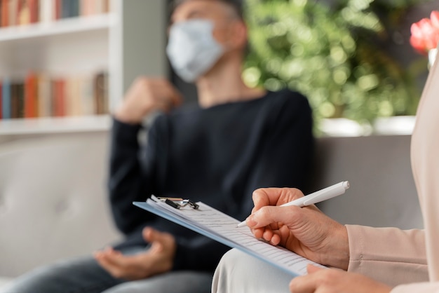 Terapeuta mujer tomando notas del hombre en la oficina de terapia con máscara