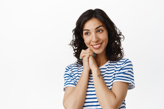 Ternura y bienestar. Hermosa chica morena mirando tiernamente a un lado, tomándose de la mano cerca de la cara romántica, sonriendo, admirando algo lindo, de pie contra el fondo blanco.