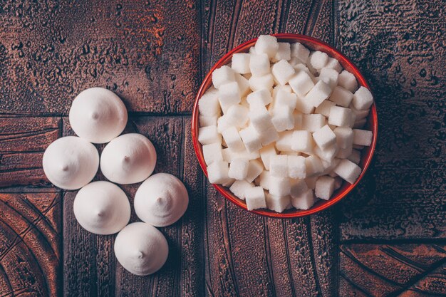 Terrones de azúcar blanco en un tazón con caramelos planos sobre una mesa de madera oscura.