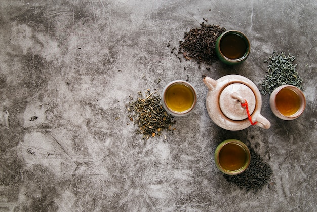 Tetera de cerámica rodeada de hierbas secas y tazas de té sobre fondo de hormigón