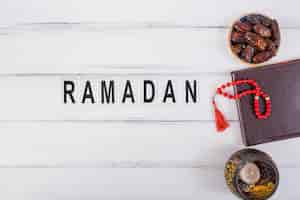 Foto gratuita texto de ramadán con cuenco de dátiles jugosos; diario y cuentas rojas de oración en mesa blanca