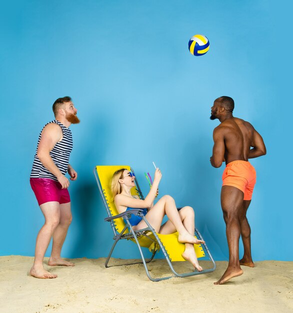 Tiempo de actividad. Amigos felices toman selfie, jugando voleibol sobre fondo azul de estudio. Concepto de emociones humanas, expresión facial, vacaciones de verano o fin de semana. Frío, verano, mar, océano.