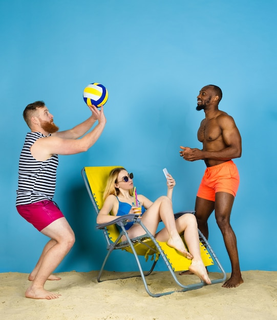 Tiempo de actividad. Amigos felices toman selfie, jugando voleibol sobre fondo azul de estudio. Concepto de emociones humanas, expresión facial, vacaciones de verano o fin de semana. Frío, verano, mar, océano.