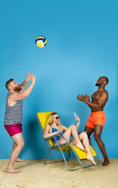 Tiempo de actividad. Amigos felices toman selfie, jugando voleibol sobre fondo azul de estudio. Concepto de emociones humanas, expresión facial, vacaciones de verano o fin de semana. Frío, verano, mar, océano.