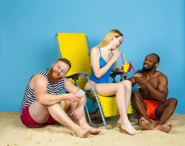 Tiempo brillante. Amigos felices descansando, bebiendo cócteles sobre fondo azul de estudio. Concepto de emociones humanas, expresión facial, vacaciones de verano o fin de semana. Frío, verano, mar, océano, alcohol.