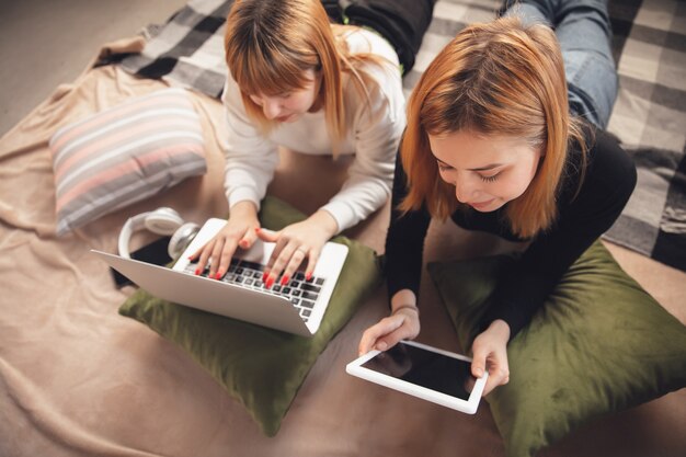 Tiempo de educación. Amigos jóvenes, mujeres que usan gadgets para ver cine, fotos, cursos en línea, tomarse selfies o vlogs, ir de compras. Dos niñas caucásicas en casa usando laptop, tableta, teléfono inteligente, auriculares.