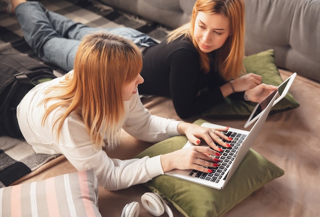 Tiempo de educación. Jóvenes amigos, mujeres que usan gadgets para ver cine, fotos, cursos en línea, tomarse selfies o vlogs, ir de compras. Dos niñas caucásicas en casa usando laptop, tableta, teléfono inteligente, auriculares.