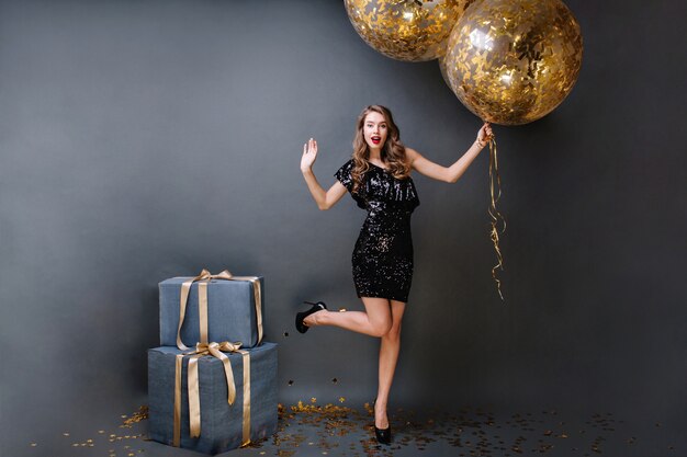 Tiempo de fiesta brillante hermosa joven en vestido de lujo negro, tacones, con cabello largo y rizado morena sosteniendo grandes globos llenos de oropel. Regalos, fiesta de cumpleaños.