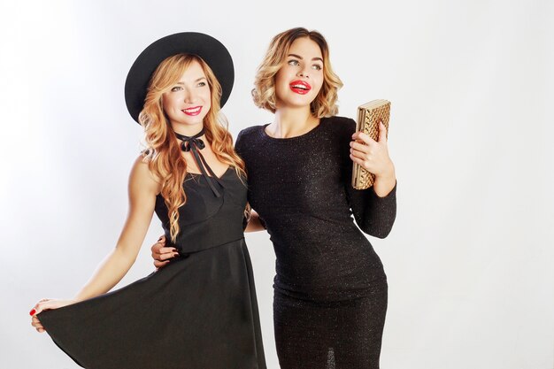 Tiempo de fiesta de dos mejores amigas, mujeres rubias en elegante vestido de cóctel negro posando en el estudio