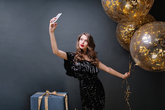 Tiempo de fiesta de mujer joven atractiva en vestido de lujo negro, con pelo largo y rizado morena haciendo selfie con globos grandes llenos de oropel. Presenta, celebrando, moderno.