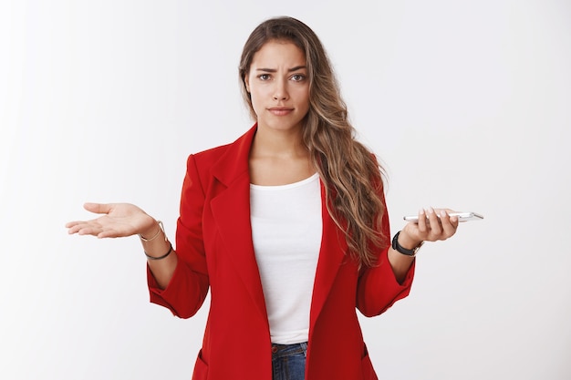 Tienes algún problema. Cuestionado molesto atractivo confiado joven caucásico mujer de pelo rizado encogiéndose de hombros consternación insegura sosteniendo las manos del teléfono inteligente extendidas hacia los lados, no puede entender lo que la persona quiere