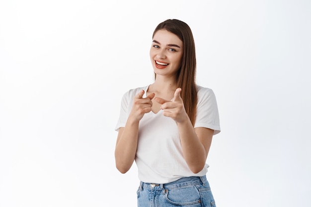 Tienes esto, únete a nosotros. Sonriente mujer asertiva apuntando al frente, invitando a trabajar para la empresa o al evento, alabando el buen trabajo, el gesto bien hecho, de pie sobre una pared blanca