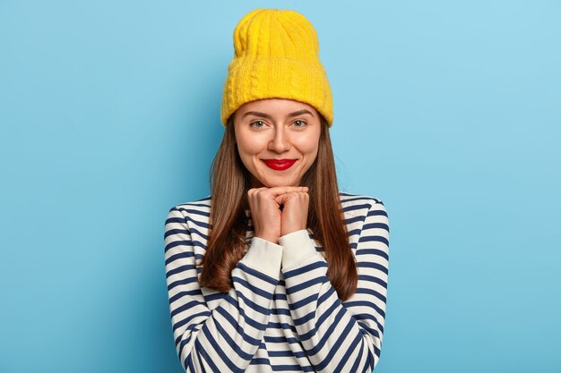 Tierna buena mujer de pelo oscuro está contra el fondo azul con sombrero amarillo y suéter a rayas, lleva lápiz labial rojo