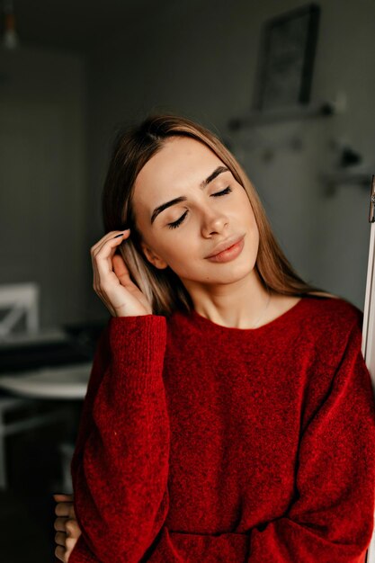 Tierna chica elegante con maquillaje desnudo vestida con suéter rojo ojos cerrados y sonriente Foto interior de mujer atractiva