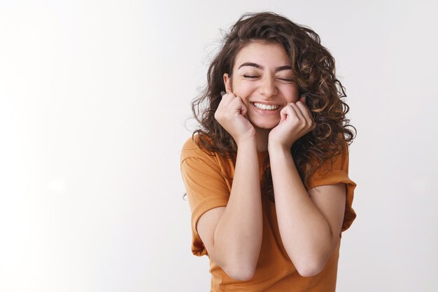 Tierna chica femenina atractiva excitada compañera de clase invitada prom sonriendo felizmente sueño hecho realidad de pie tocado apasionado ojos cerrados con alegría animando prensa manos mejillas, fondo blanco