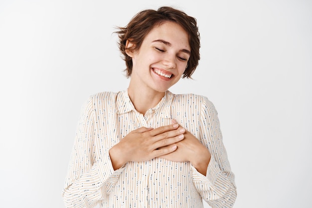 Tierna chica morena en blusa soñando con algo romántico cerrar los ojos y sonreír con las manos en el corazón de pie sobre fondo blanco.