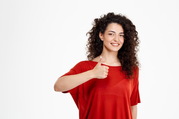 Tierna chica rizada sonriendo y señalando bien firmar sobre pared blanca