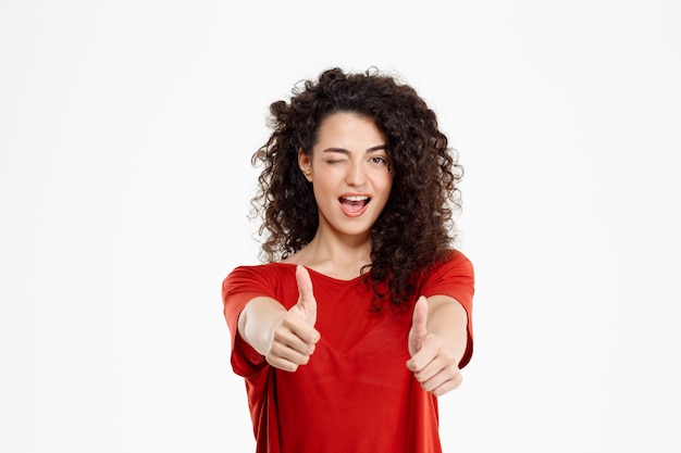 Tierna chica rizada sonriendo y señalando bien firmar sobre pared blanca