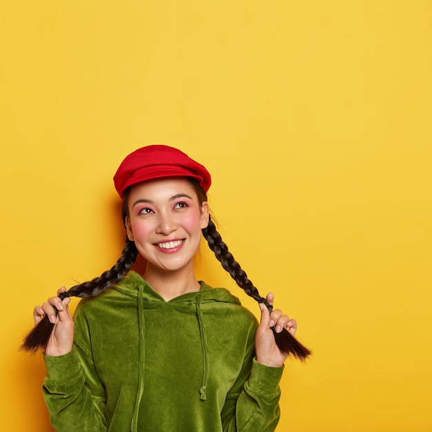 Foto gratuita la tierna y fascinada joven coreana muestra largas trenzas, tiene una expresión pensativa de ensueño, vestida con ropa elegante y vívida