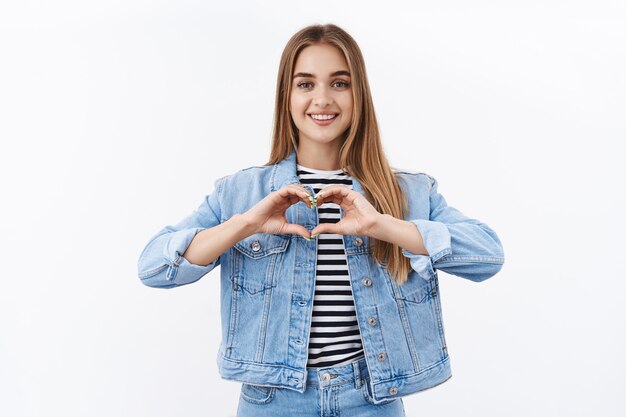 Tierna joven enamorada confiesa simpatía, muestra gesto de corazón y sonríe, mira a la cámara romántica, adora algo, como producto o persona, de pie en la pared blanca alegre