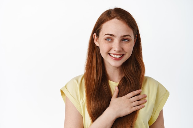 La tierna y linda modelo femenina pelirroja sosteniendo la mano en el corazón se siente romántica, soñando despierto en blanco