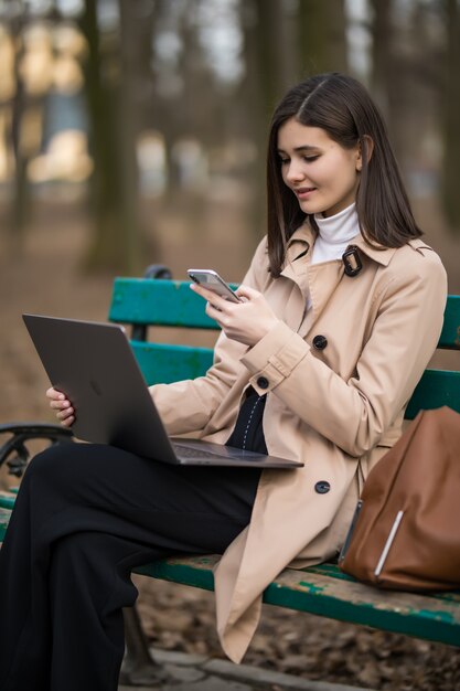 Tierna morena modelo trabaja en la computadora portátil y el teléfono afuera en el parque