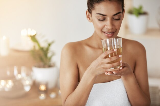 Tierna mujer africana sonriente sosteniendo un vaso con té descansando en el salón de spa