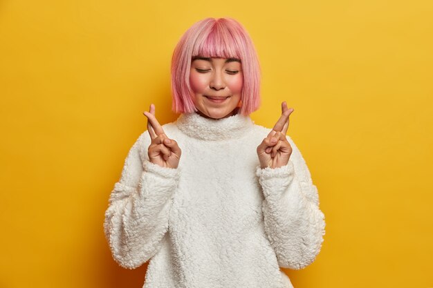 Tierna mujer asiática sonriente con cabello rosado, cierra los ojos y cruza los dedos para la buena suerte, pide deseos, tiene fe, quiere lograr el éxito