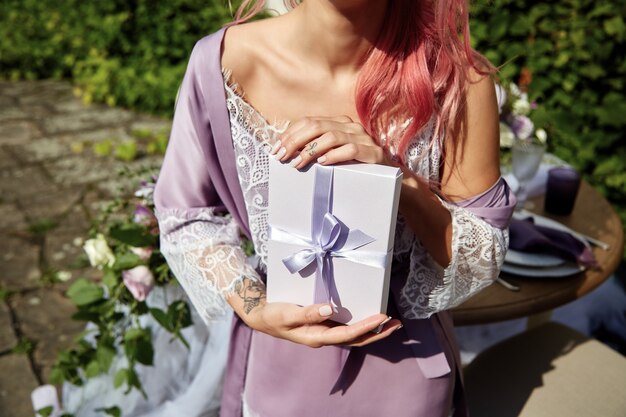 Tierna mujer con cabello rosado posa en bata violeta con caja blanca presente
