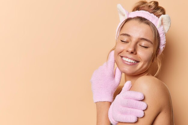 Tierna mujer joven y alegre disfruta de una piel suave y saludable después de los procedimientos de spa usa diadema y guantes de ducha mantiene los ojos cerrados usa diadema para hombros desnudos contra el espacio de copia de fondo marrón