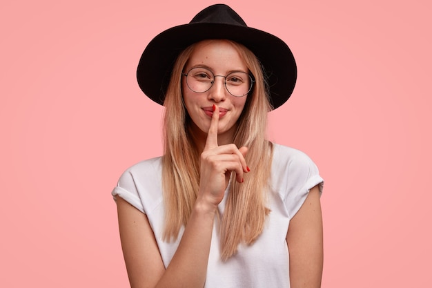 Tierna mujer joven hermosa con expresión complacida, hace gesto de silencio, mira feliz y dice shh