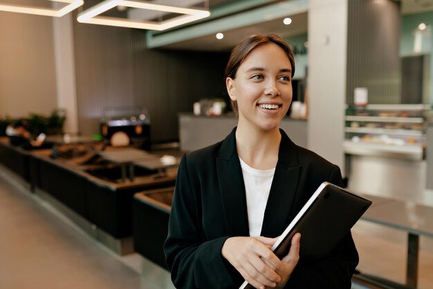 Tierna mujer de negocios sonriente en elegante blusa y chaqueta negra sosteniendo una computadora portátil y tomando café en la oficina La mujer moderna está trabajando en la oficina y esperando a colegas universitarios
