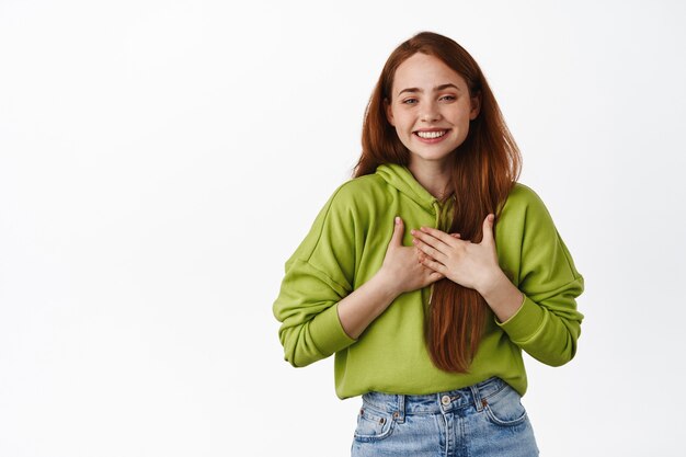 Tierna niña sonriente con cabello rojo, tomados de la mano en el pecho y riendo complacida, sentirse agradecida, decir gracias, apreciar la ayuda, de pie sobre blanco.