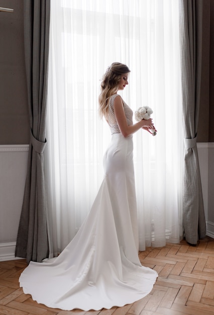 Tierna novia rubia caucásica en elegante vestido con ramo de novia blanco está de pie cerca de la ventana