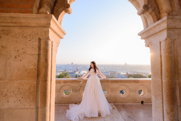 Tierna novia vestida con un elegante vestido de novia está de pie en el balcón de un antiguo edificio de piedra